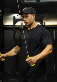 Άνδρας με μαύρη αθλητική φόρμα και καπέλο που χρησιμοποιεί μηχάνημα γυμναστηρίου με καλώδια και κίτρινες λαβές, εστιάζοντας στην προπόνηση δύναμης.