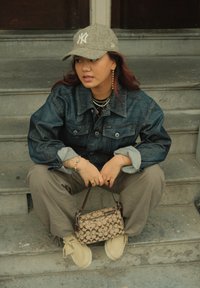 Denimjakke, grå caps, lagdelte halskjeder, beige veske med monogrammønster, grå bukser og lette sko. Sitter på steintrapper.