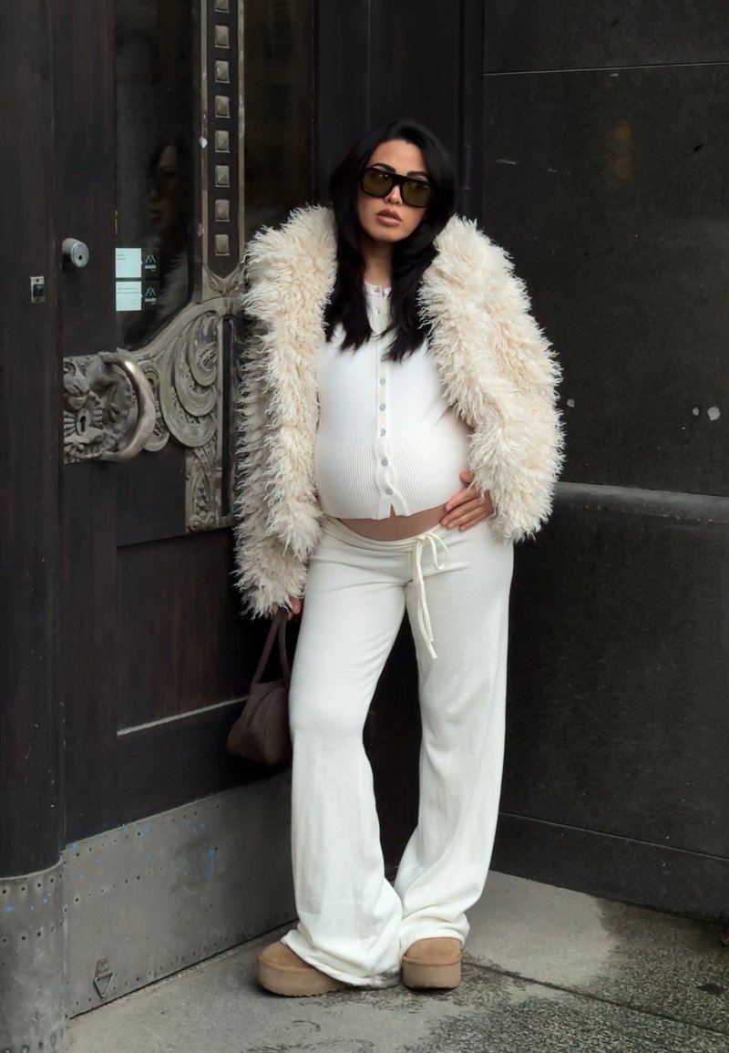 Femme enceinte portant un pantalon blanc, un haut blanc boutonné, une veste beige duveteuse, des lunettes de soleil et des chaussures marron, debout près d'une porte sombre ornée.