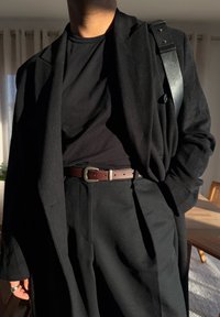 Μαύρο μάλλινο παλτό σε στρώσεις πάνω από ένα μαύρο πουκάμισο, σε συνδυασμό με προσαρμοσμένο μαύρο παντελόνι και καφέ δερμάτινη ζώνη. Λείες υφές και ελάχιστες λεπτομέρειες.