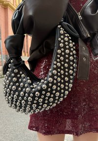 Schwarze Lederhandtasche mit silbernen Nieten, abgerundeter Form und einem geknoteten Griff. Platziert neben einem burgunderroten, paillettenbesetzten Kleid.