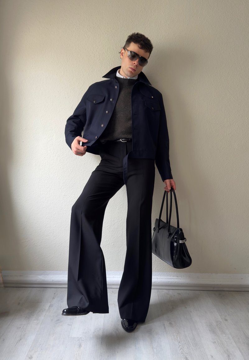 Person, die dunkle, weit geschnittene Hosen, eine marineblaue Jacke, einen grauen Pullover und eine Sonnenbrille trägt, hält eine schwarze Handtasche und posiert vor einer beigen Wand auf einem Holzboden.