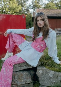 Salopette rosa a fantasia indossata sopra una maglietta azzurro chiaro, abbinata a stivaletti bianchi alla caviglia. Sullo sfondo, un ambiente naturale e un contenitore rosso.