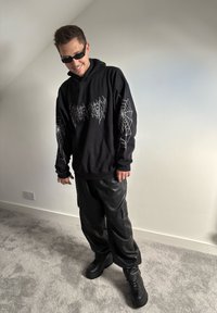 Czarna bluza z kapturem z białymi grafikami w kształcie pajęczej sieci i napisem, zestawiona z błyszczącymi czarnymi spodniami cargo i platformowymi czarnymi sneakersami. Gładkie tekstury.