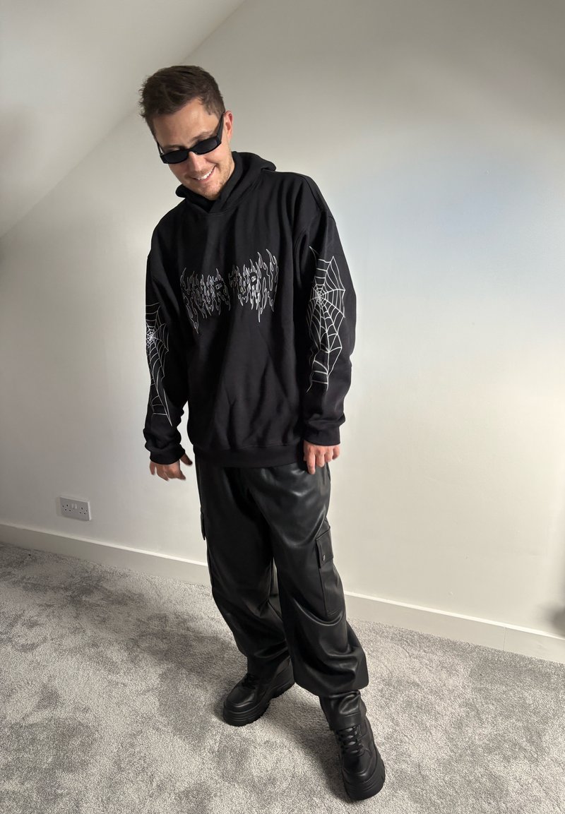 Schwarzer Hoodie mit weißen Spinnennetzgrafiken und Text, kombiniert mit glänzenden schwarzen Cargo-Hosen und schwarzen Plateauschuhen. Glatte Texturen.
