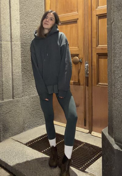 Sudadera gris oscuro de gran tamaño con bolsillo canguro, combinada con leggings verde oscuro. Botines marrones con calcetines blancos, de pie frente a una puerta de madera.