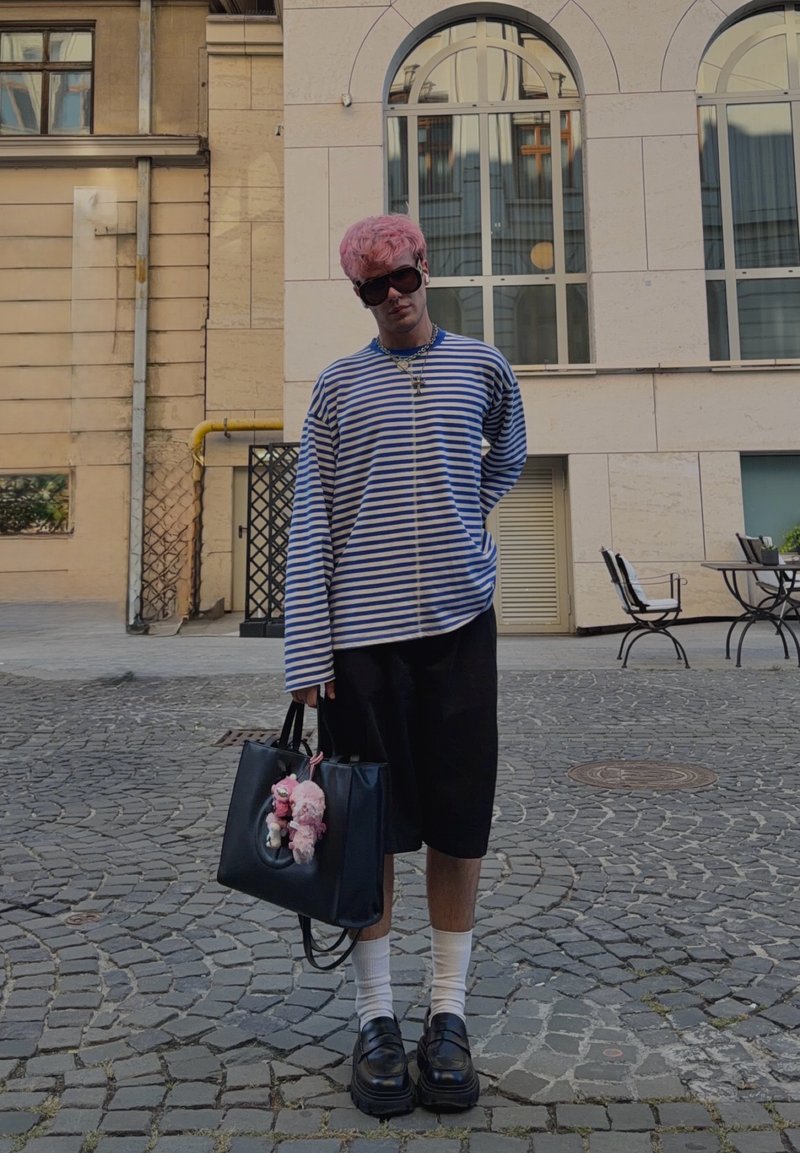 Personne aux cheveux roses et lunettes de soleil se tient dans une rue pavée, portant une chemise rayée, un short noir, des chaussettes blanches, des chaussures noires, tenant un sac noir avec des peluches.