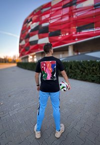 Μαύρη μπλούζα T-shirt με πολύχρωμο αφηρημένο σχέδιο στην πλάτη, φορεμένη με ανοιχτό μπλε παντελόνι φόρμας, κρατώντας μια μπάλα ποδοσφαίρου, outdoors κοντά σε ένα στάδιο.