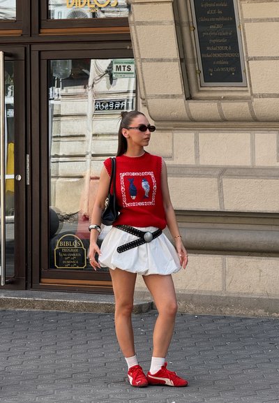Top sin mangas de punto rojo con diseño de pez bordado, falda blanca acampanada, cinturón negro con hebilla circular plateada y zapatillas deportivas rojas.