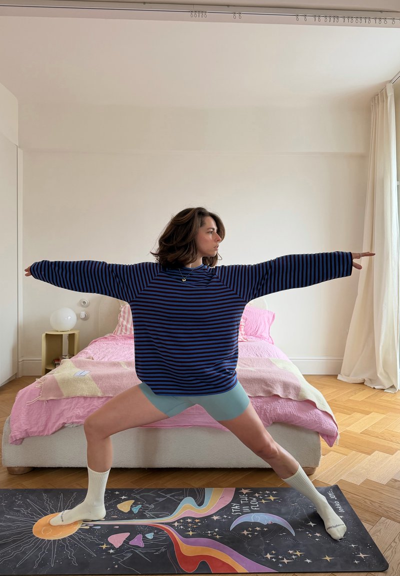 Person i randig tröja och shorts som gör yogapositionen Krigare II på en färgglad matta i ett minimalistiskt sovrum med rosa säng och trägolv.