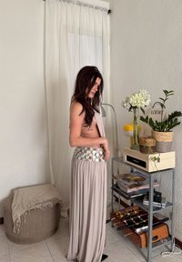 Femme aux longs cheveux foncés portant une jupe fluide ornée de sequins, debout à côté d'une étagère métallique contenant des plantes, des livres et une table de baby-foot à l'intérieur.