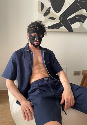 Camisa de manga corta azul marino con ribete blanco, combinada con pantalones cortos a juego. Cara cubierta con una mascarilla negra, sentado en un puff de color claro.
