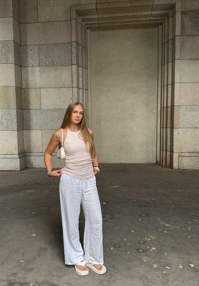 Top ajustado de color rosa claro combinado con pantalones sueltos de lino blanco. Los accesorios incluyen un pequeño bolso blanco y una pulsera. Fondo de pared de piedra.