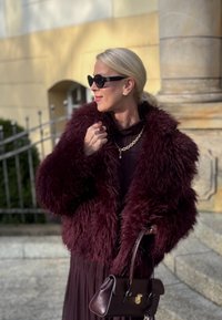 Burgundy fuskpälsjacka med voluminös textur, lager på lager över en mörk klänning, ihop med en liten brun handväska och svarta cat-eye solglasögon.