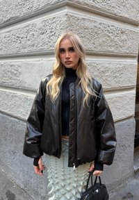 Czarna skórzana kurtka typu bomber z pikowanym wzorem, ściągaczami na rękawach i zamkiem błyskawicznym; zestawiona z teksturowaną jasną spódnicą.