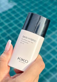 Fondotinta Skin Hybrid Blurring di KIKO Milano SPF 30 in una boccetta beige opaco con un tappo nero, tenuto davanti a uno sfondo di piastrelle blu.