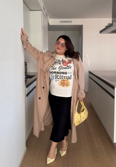 Larga gabardina beige, camiseta gráfica blanca con estampados de comida, falda midi negra, bolso amarillo y tacones puntiagudos de un amarillo claro.