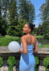 Robe sans bretelles bleu clair, tissu lisse, silhouette ajustée. Tenant un ballon blanc, se tenant près d'une rambarde en pierre avec un fond de verdure.