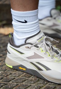 Las zapatillas Reebok presentan una parte superior blanca con acentos grises, malla texturizada, detalles en verde lima y suela de Vibram. Se llevan con calcetines blancos de Nike.
