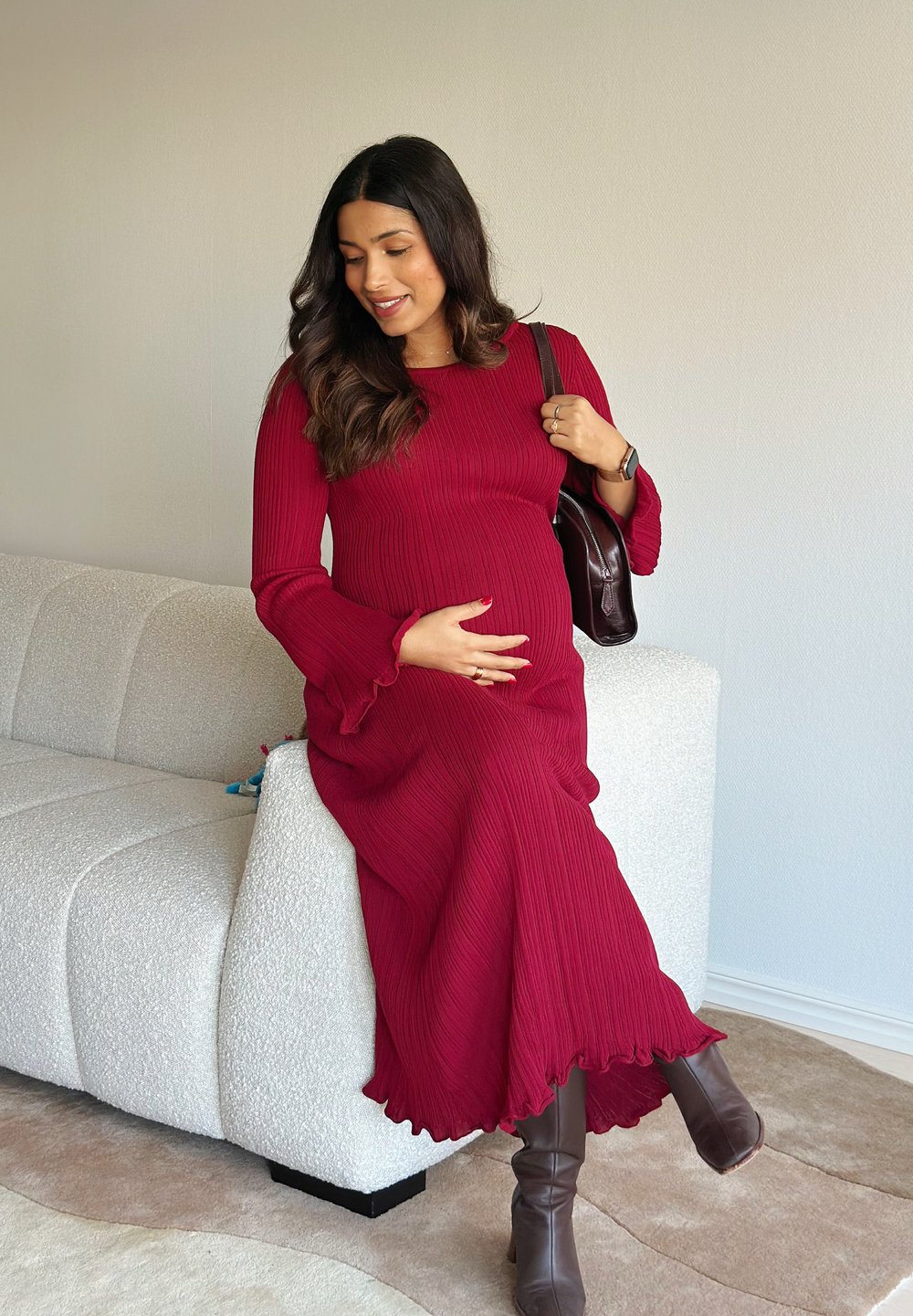Lange, geribbelde wijnrode jurk met uitlopende mouwen en ruches aan de zoom. Gecombineerd met bruine kniehoge laarzen. Bruine handtas rust op de schouder.