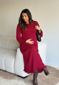 Lange, geribbelde wijnrode jurk met uitlopende mouwen en ruches aan de zoom. Gecombineerd met bruine kniehoge laarzen. Bruine handtas rust op de schouder.