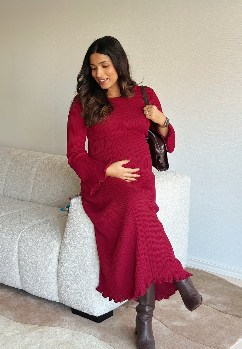 Robe longue bordeaux à côtes avec manches évasées et ourlet à volants. Portée avec des bottes marron montant jusqu'aux genoux. Sac à main marron reposant sur l'épaule.