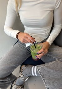 Zeleno pijačo v prozorni steklenici s črno slamico; obrabljeni sivi kavbojke; vzorčaste obutve; svetla dolga majica s dolgimi rokavi; temno nohtni lak na prstih.