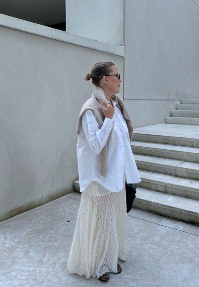 Camisa blanca de gran tamaño con botones, suéter de punto beige drapeado sobre los hombros, falda larga y ligera de color crema, bolso negro pequeño, zapatos de tacón cuadrado.