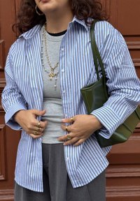 Chemise à rayures bleues et blanches boutonnée sur un haut gris à manches longues, associée à un pantalon gris et un sac à main vert. Plusieurs bagues en or.