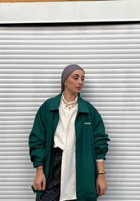 Casaco verde leve sobre uma camisa branca de botões, combinado com uma saia preta com padrão. Os acessórios incluem joias douradas grossas e um lenço cinza na cabeça.