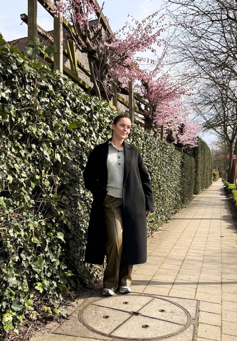 Woman in black coat and beige pants leaning against a green ivy-covered wall, pink cherry blossoms above, on a paved sidewalk.