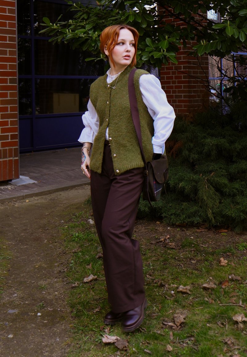 Persona con capelli rossi che indossa un gilet verde oliva, una camicetta bianca e pantaloni marroni, sta all'aperto vicino a un muro di mattoni e a un cespuglio foglioso, guardando di lato.