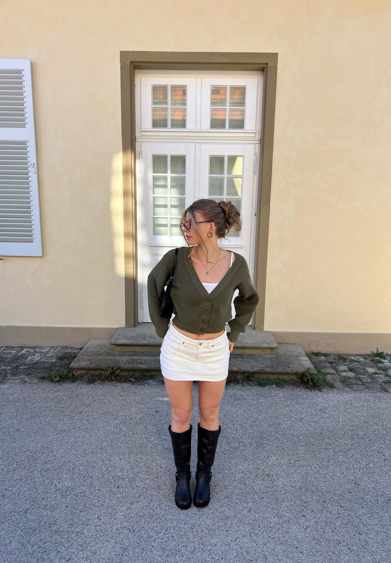 Femme avec des lunettes portant un cardigan vert, une mini-jupe blanche et des bottes noires, debout devant une porte blanche avec panneaux en verre.