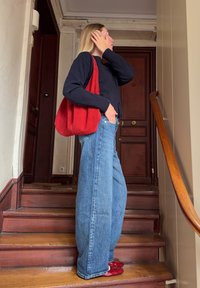 Γυναίκα με μακριά ξανθά μαλλιά, που φοράει μαύρο πουλόβερ, φαρδιά μπλε τζιν, κόκκινα παπούτσια και κόκκινη τσάντα, στέκεται σε εσωτερική ξύλινη σκάλα δίπλα σε καφέ πόρτες.