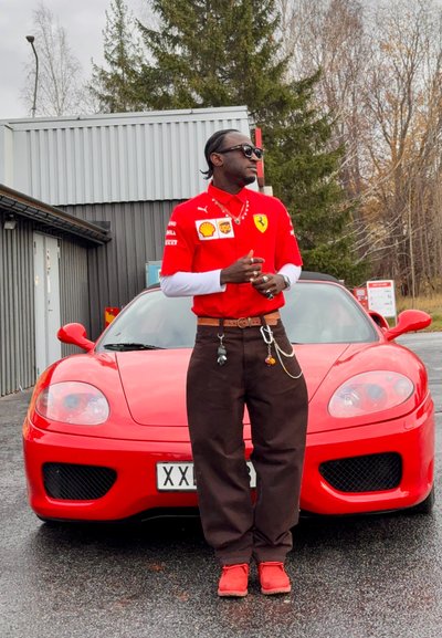 Coche deportivo Ferrari rojo con diseño aerodinámico y parrilla oscura, estacionado sobre el asfalto. Persona con camiseta roja de marca con detalles blancos y pantalones negros está cerca.