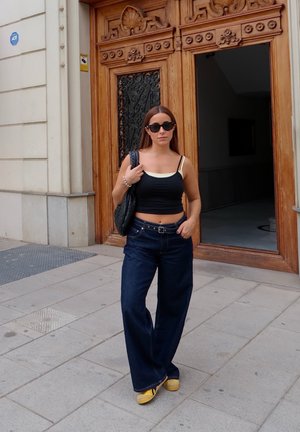 Top corto negro con ribete crema, jeans oscuros de pierna ancha, bolso negro y zapatos amarillos. De pie junto a una puerta de madera.