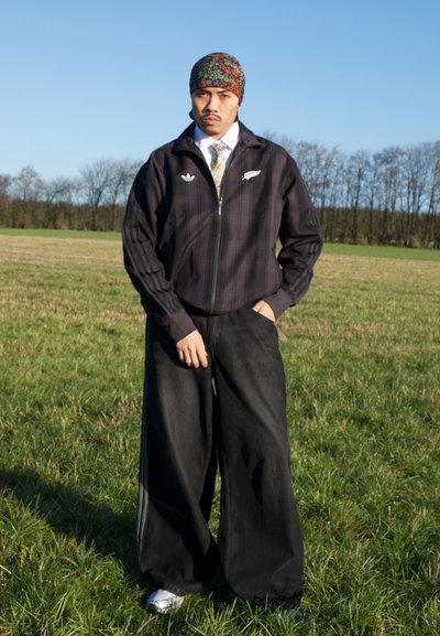 Hombre de pie en un campo de hierba vistiendo un pañuelo colorido con estampado en la cabeza, chaqueta oscura con cremallera, camisa blanca con corbata, pantalones anchos negros y zapatillas.