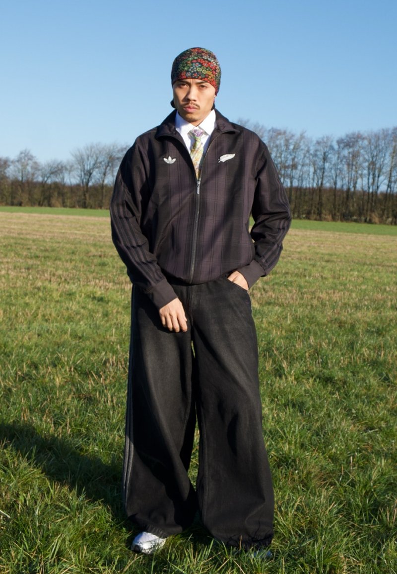 Homme debout dans un champ herbeux, portant un foulard coloré à motifs, une veste zippée sombre, une chemise blanche avec une cravate, un pantalon large noir et des baskets.
