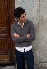 Homme en pull gris à torsades porté sur une chemise blanche, debout les bras croisés contre un mur en pierre et une porte en bois.