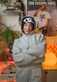 Sweat à capuche gris zippé et pantalon de survêtement, bonnet noir et blanc avec logo, superposés dans un cadre intérieur vibrant avec des plantes et de la décoration.