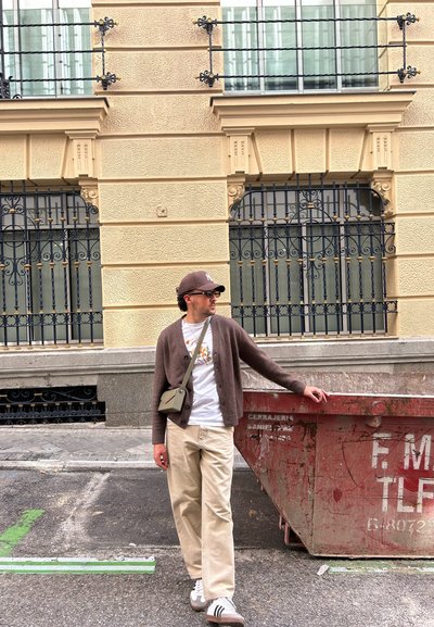 Conjunto informal que incluye un cárdigan marrón, una camiseta gráfica, pantalones beiges y zapatillas a rayas gris-blancas. Se incluye un pequeño bolso bandolera.