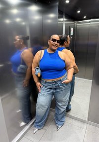 Modrý pruhovaný crop top, světle modré kalhoty s širokými nohavicemi. Stříbrné špičaté boty. Doplňky zahrnují malou modrou tašku a sluneční brýle. Odraz v zrcadle.