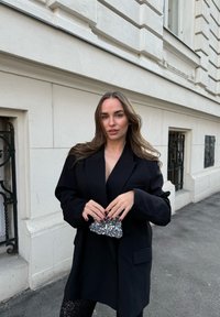 Fekete, oversized blézer mély V-nyakkal, fényes mintás leggingszel párosítva. Csillogó clutch táska a kezében. Semleges h�áttér.