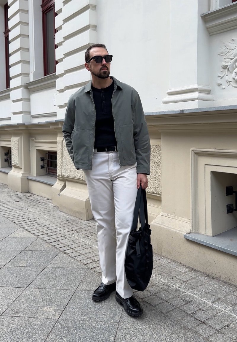 Homme portant des lunettes de soleil, une veste grise, un pantalon blanc, des chaussures noires, tenant un sac noir, marchant dans une rue de la ville près d'un bâtiment beige.