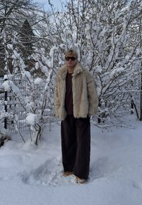 Manteau en fausse fourrure beige, pantalon long marron, chemise marron et bottes couleur sable. Debout dans la neige, avec un arbre enneigé en arrière-plan.