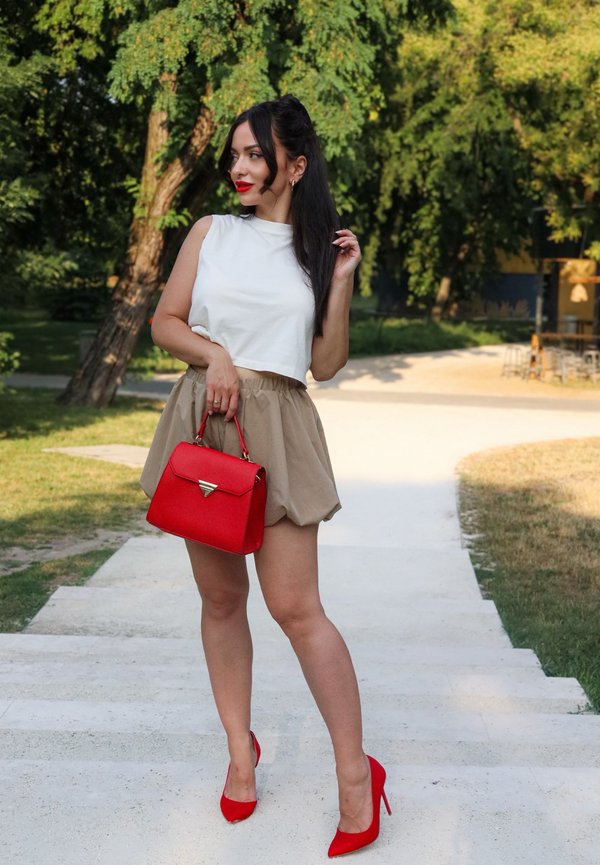 Frau in weißem ärmellosen Oberteil, beigem Rock, roten High Heels und einer roten Handtasche, die auf Außentreppen steht mit Bäumen im Hintergrund.