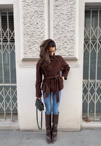 Femme portant une veste marron ceinturée, un jean bleu et de longues bottes marron, debout dans la rue, tenant un sac à main noir et regardant vers le bas.