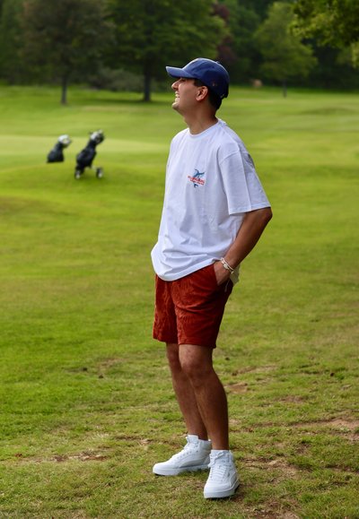 Camisa blanca de manga corta con logo gráfico, pantalones cortos texturizados en marrón y zapatillas blancas. El fondo presenta césped verde y bolsas de golf.