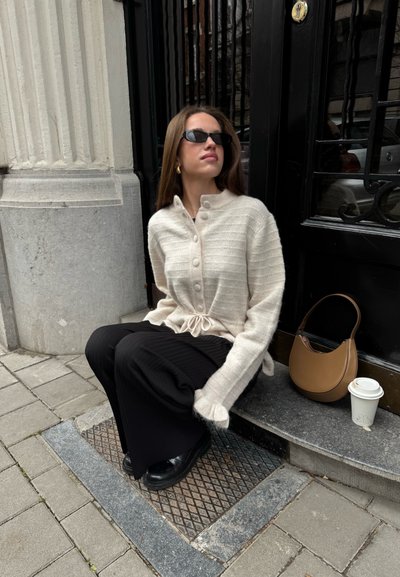 Cardigan beige claro con botones, textura acanalada y lazo a juego en la cintura, combinado con pantalones de pierna ancha negros y zapatos negros. Bolso marrón y taza de café.