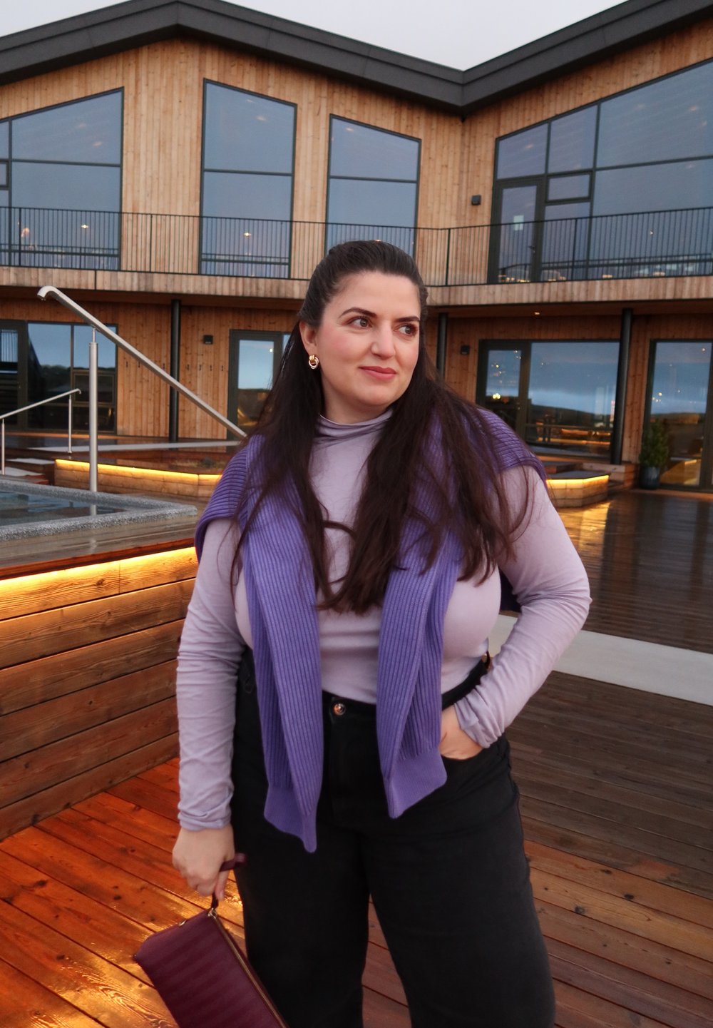 Lila gerippter Pullover über einem enganliegenden helllila Oberteil, kombiniert mit hoch taillierten schwarzen Hosen. Hält eine bordeauxrote Clutch. Hintergrund aus Holzdeck.